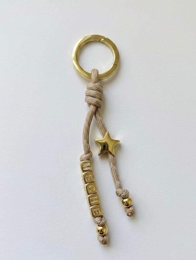 Keychain / bag charm with gold ring, star, and text charm on beige cord against a white background