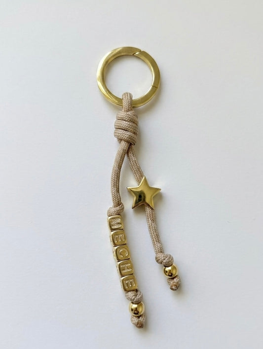 Keychain / bag charm with gold ring, star, and text charm on beige cord against a white background